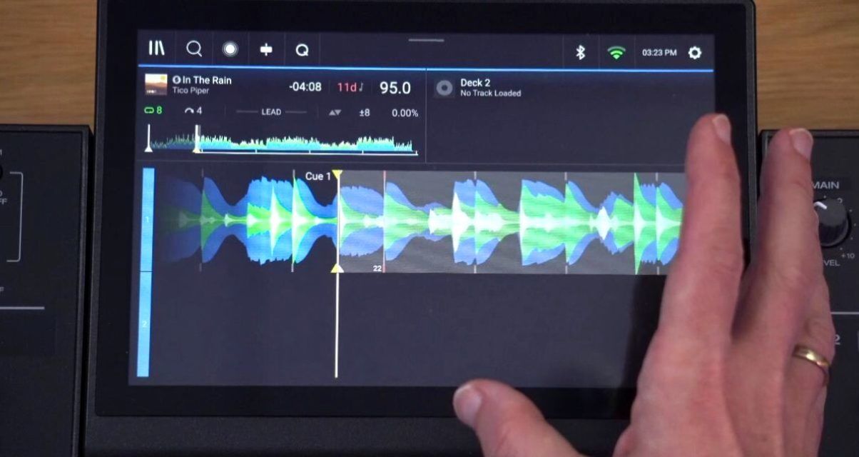 Top-down view of the Denon DJ Prime 4+ screen laying on a wooden desk, with the hand of a white man gesturing towards the blue white and green track waveform