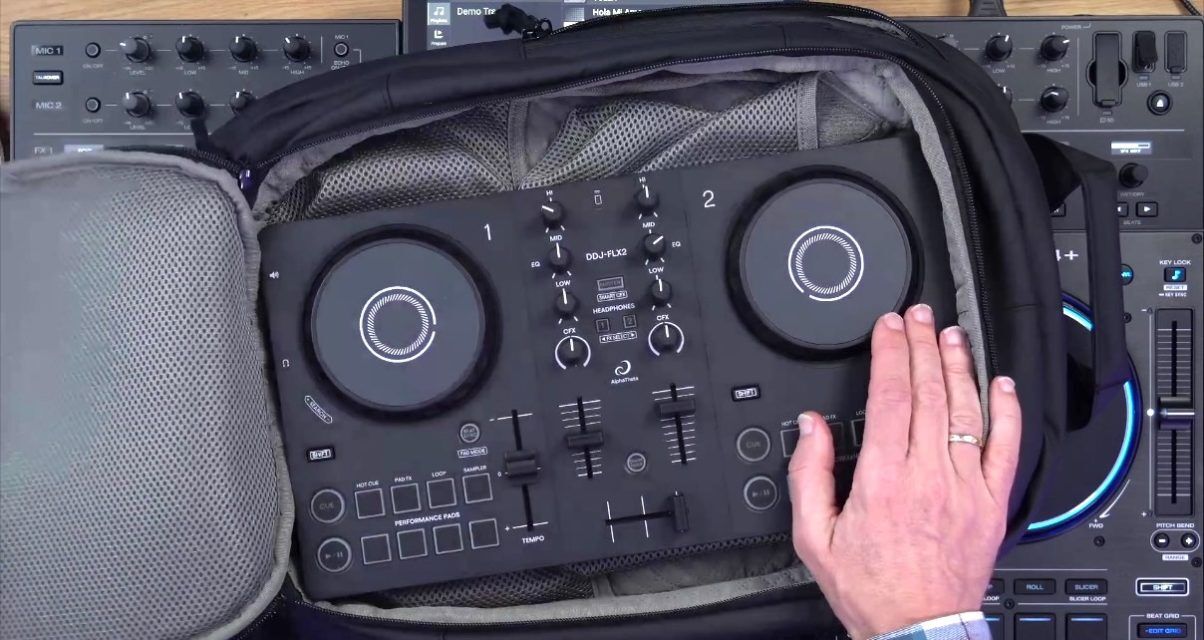 Top-down view of a black DDJ-FLX2 DJ controller sitting in a bag, being held my a white person's right hand. Underneath everything is a dark grey Denon DJ Prime 4+ unit.