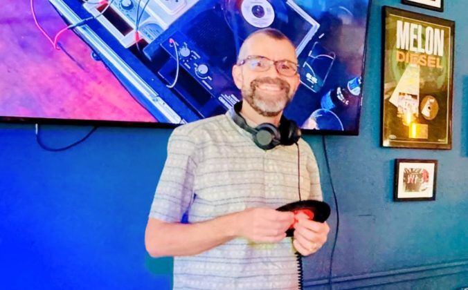 Digital DJ Tips founder Phil Morse smiling in front of a large TV displaying a vinyl DJ set-up. Phil is a white man with glasses and a beard, wearing black headphones around his neck and a light striped t-shirt. He's looking at the camera and holding a seven-inch record with both hands.