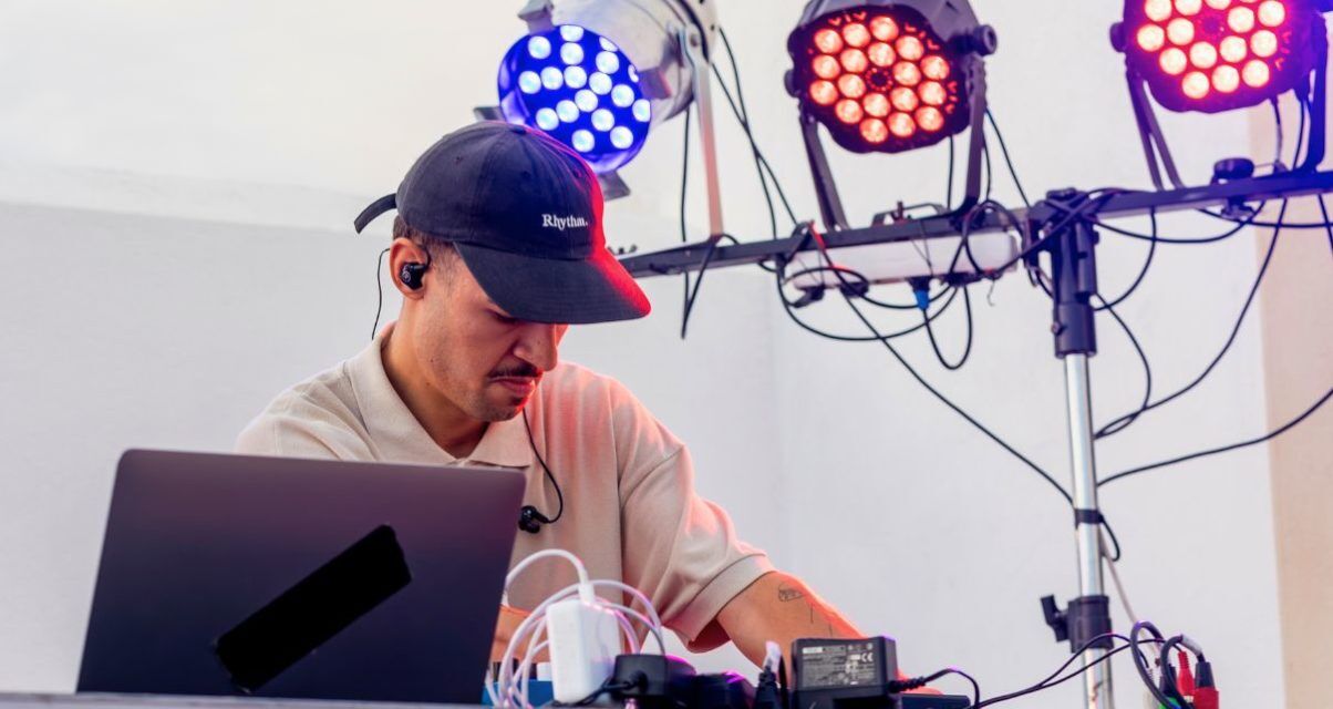 A DJ with a hat, mustache, and IEMs tweaks his DJ gear. In the background are three different coloured stage lights.