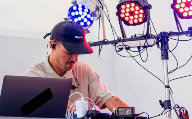 A DJ with a hat, mustache, and IEMs tweaks his DJ gear. In the background are three different coloured stage lights.