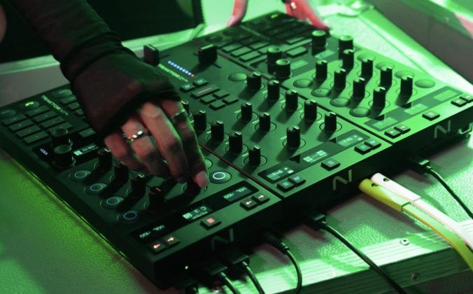 A DJ stands behind three modular Traktor units, tweaking a knob on the mixer. The photo is bathed in green lighting.