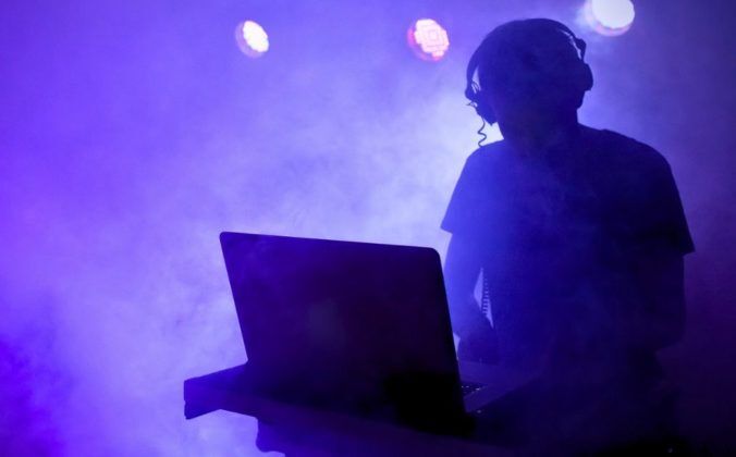 The silhouettze of a DJ stands behind a laptop. Behind the DJ are several stage lights, and the whole scene is bathed in a purple smoke machine haze.