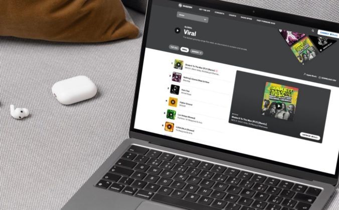 A silver laptop resting on a cream-coloured couch, next to a pair of wireless ear buds. On the laptop screen is Shazam's Viral charts page.