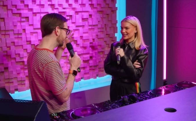 Two people holding microphones stand behind a pro DJ set-up in a bright pink and blue studio. The white man on the left has short brown hair, dark glasses, a short beard, and is wearing a red and white striped t-shirt. On the right is a white woman with blonde hair, wearing a black leather jacket. They are both smiling while talking to each other.
