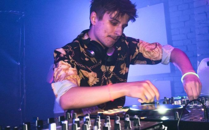 A young male DJ with short brown hair stands behind a professional DJ booth in a nightclub. He's looking down at the gear and tweaking various knobs.