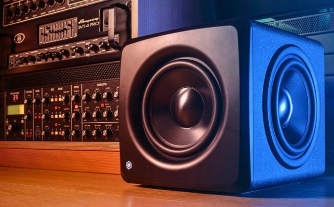 A single black iLoud Compact Studio Subwoofer sits on a wooden floor next to a rack of audio equipment.