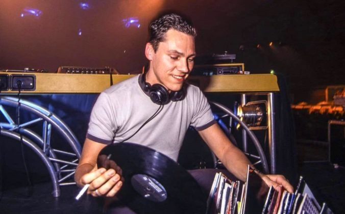 A late 90s Tiësto, smiling and facing the camera, knelt down behind the DJ booth. He's holding a cigarette and vinyl record while sorting through his collection.