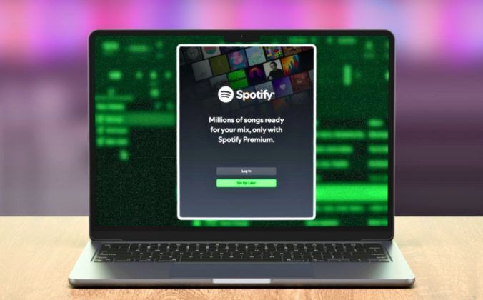A silver laptop sitting on a light wooden desk displays a Spotify login screen. In the background is a blurry DJ software screen.