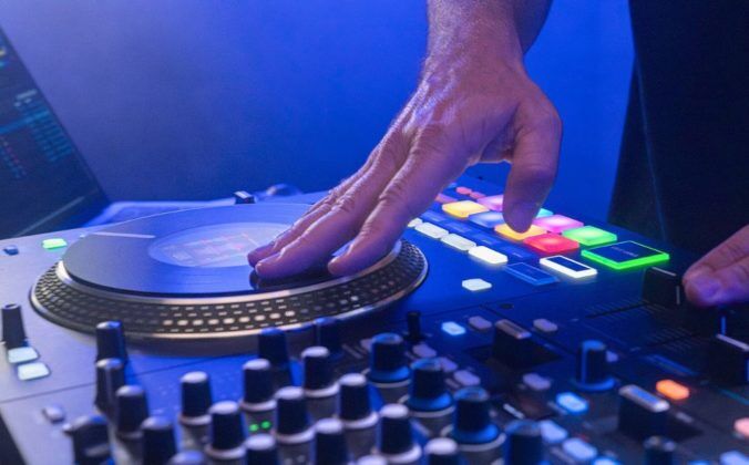 Angled view of a DJ touching the platters on a Rane Performer controller. In the background you can see a laptop displaying Serato DJ Pro software. The whole photo is lit in dark blue lighting.