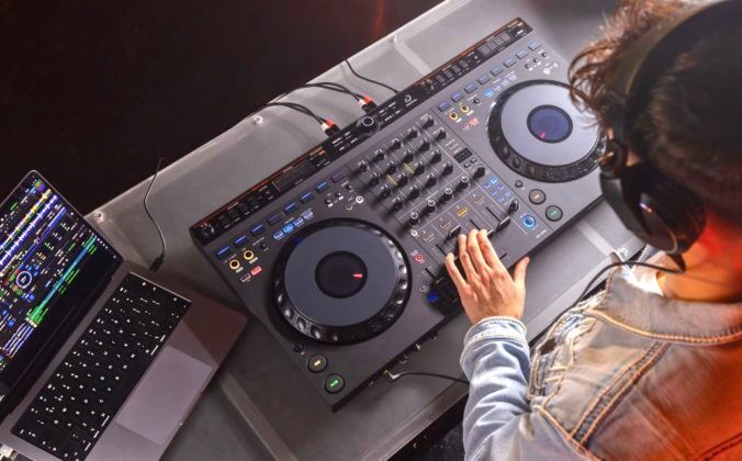 Birds eye view of a masculine-presenting brunette person standing behind a DJ booth, wearing black headphones and a denim jacket. They're looking down at an AlphaTheta DDJ-GRV6 controller and tweaking the volume levels. On the left is a silver MacBook Pro laptop displaying Rekordbox DJ software.