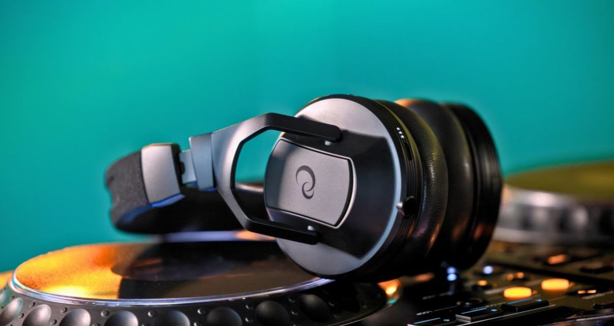 A pair of black AlphaTheta HDJ-F10 headphones laying across a CDJ-style media player. The wall in the background is emerald green.