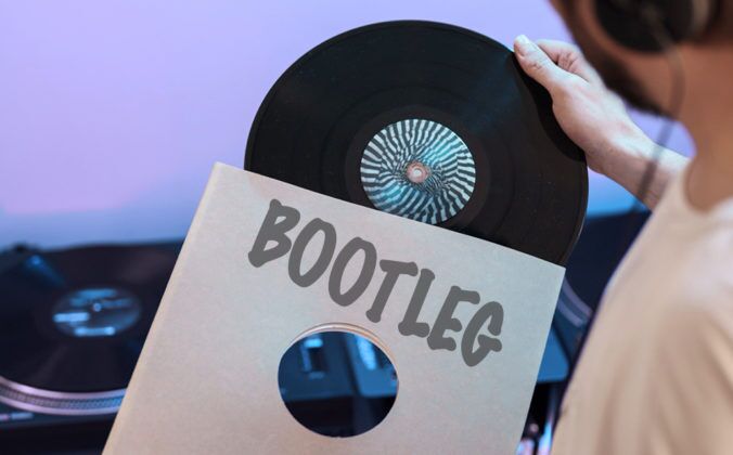 A bearded white man in his 30s stands facing a vinyl DJ set-up. He's taking a black record out of it's sleeve with his right hand. On the lightly coloured sleeve is the word "Bootleg" in big, bold letters.