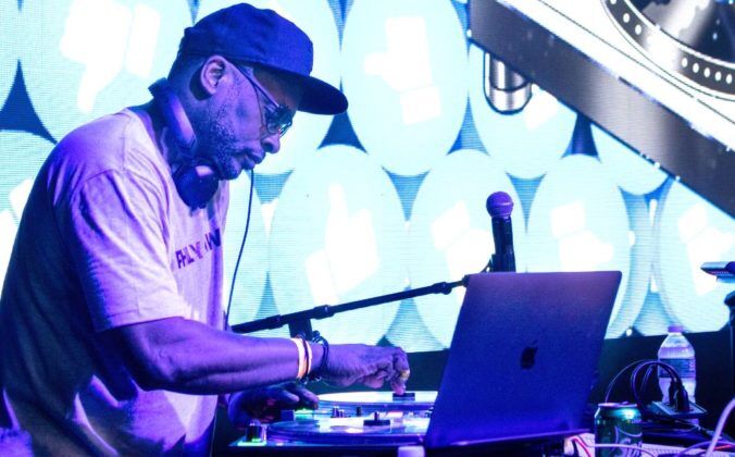 DJ Jazzy Jeff performs on stage, mixing tracks with turntables and a laptop, illuminated by vibrant blue digital graphics in the background.