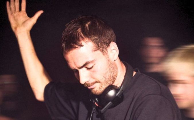 A white male-presenting DJ with short dark hair and a beard raises his hand while performing in a lively, dimly lit venue, surrounded by an energetic crowd. He is wearing a black t-shirt with a pair of black Røde NTH-50 headphones around his neck.